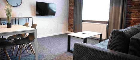 Una habitación moderna con una mesa de comedor, sillas, un sofá, una mesita de centro pequeña, un televisor de pantalla plana y un ventanal grande con cortinas.