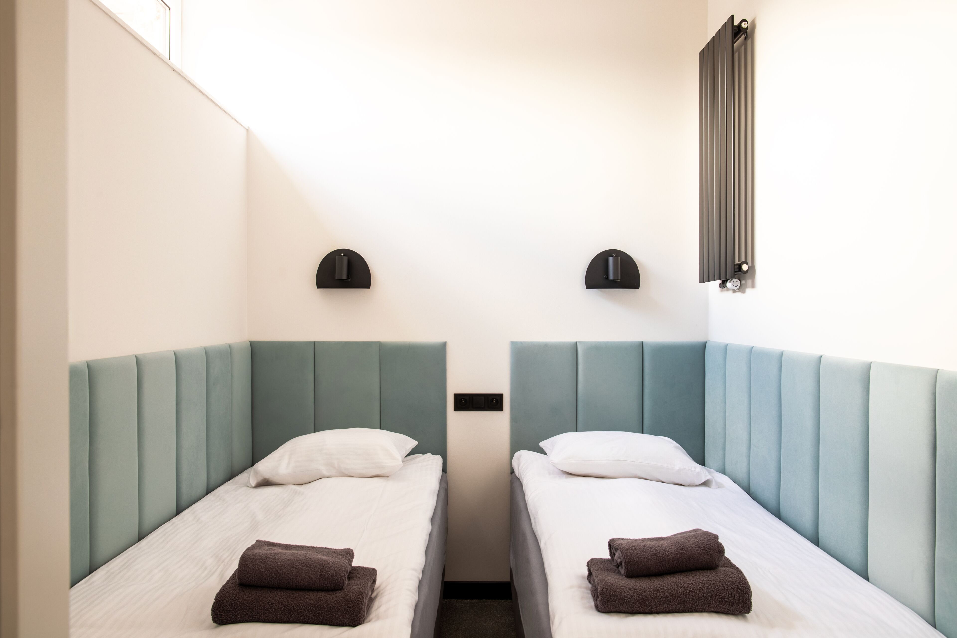 Habitación de hotel con dos camas individuales, cada una con cabecero, ventana y lámpara de pared.