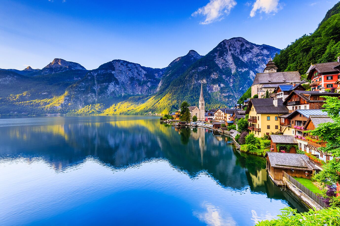 A serene lakeside town with traditional buildings, a church, and mountains in the background.