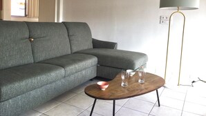 A living room with a grey sectional sofa, a wooden coffee table with two glasses and a bowl, a floor lamp, and a wall-mounted light fixture.
