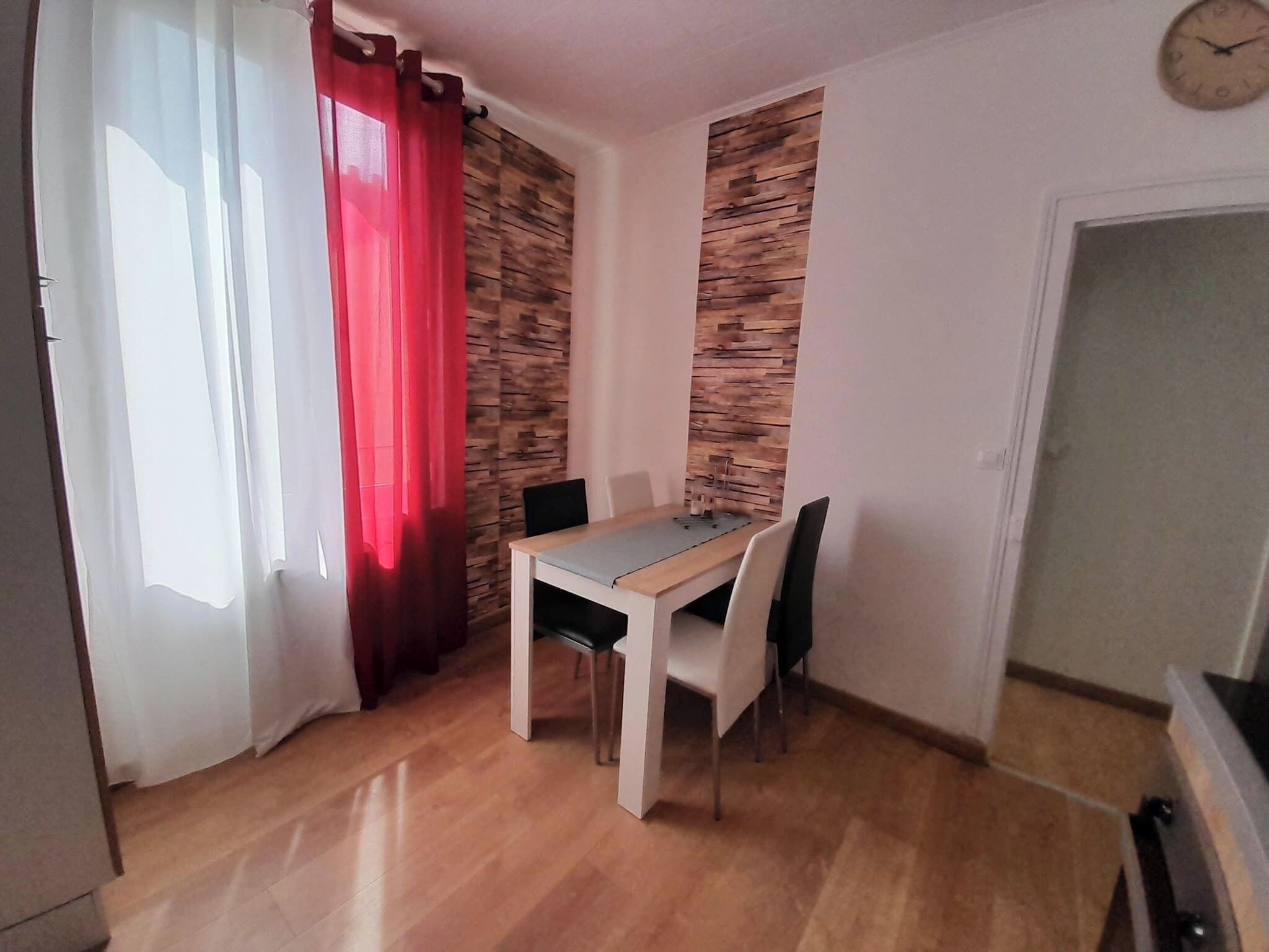 A small dining area with a wooden table and chairs, a red curtain, and a clock on the wall.