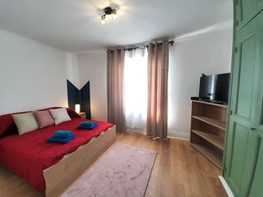 A bedroom with a wooden bed, a TV on a shelf, a nightstand, and a window with curtains. - Appart cosy-4 pers-centre ville-jardin-wifi (Châteauroux)