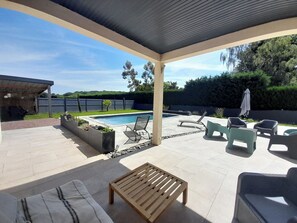 Ein überdachter Patio-Bereich mit Pool, Liegestühlen und einem Holztisch.