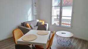 A small, bright room with a dining table, two chairs, a sofa with cushions, a round side table, and a window with a balcony view.