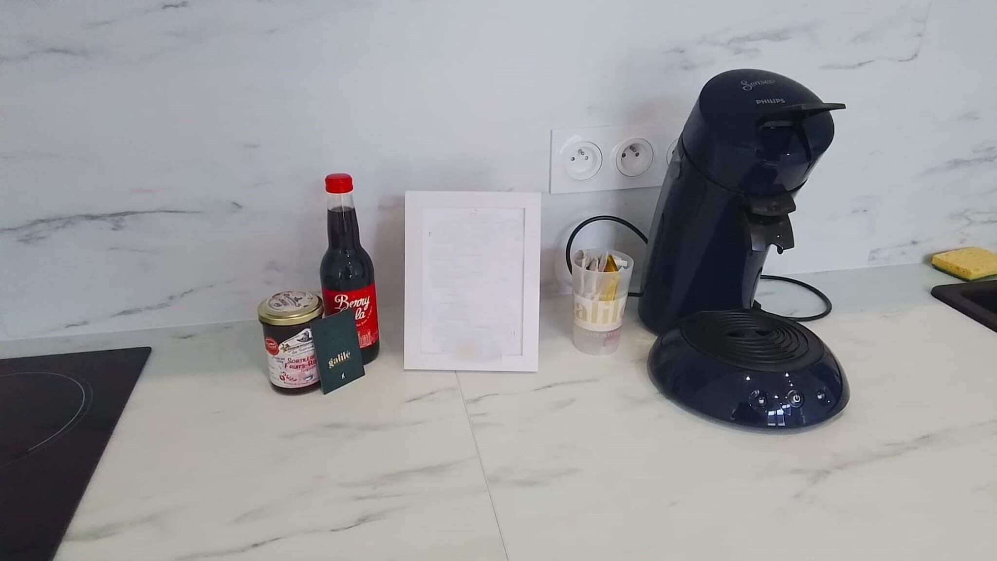 A kitchen counter with a coffee maker, a bottle of Beryl's, a jar of galette des raisins, and a framed menu.