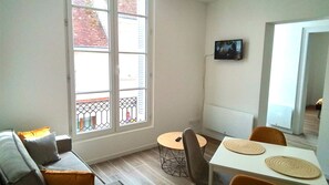 A small, bright room with a sofa, a round table, and a TV on the wall.