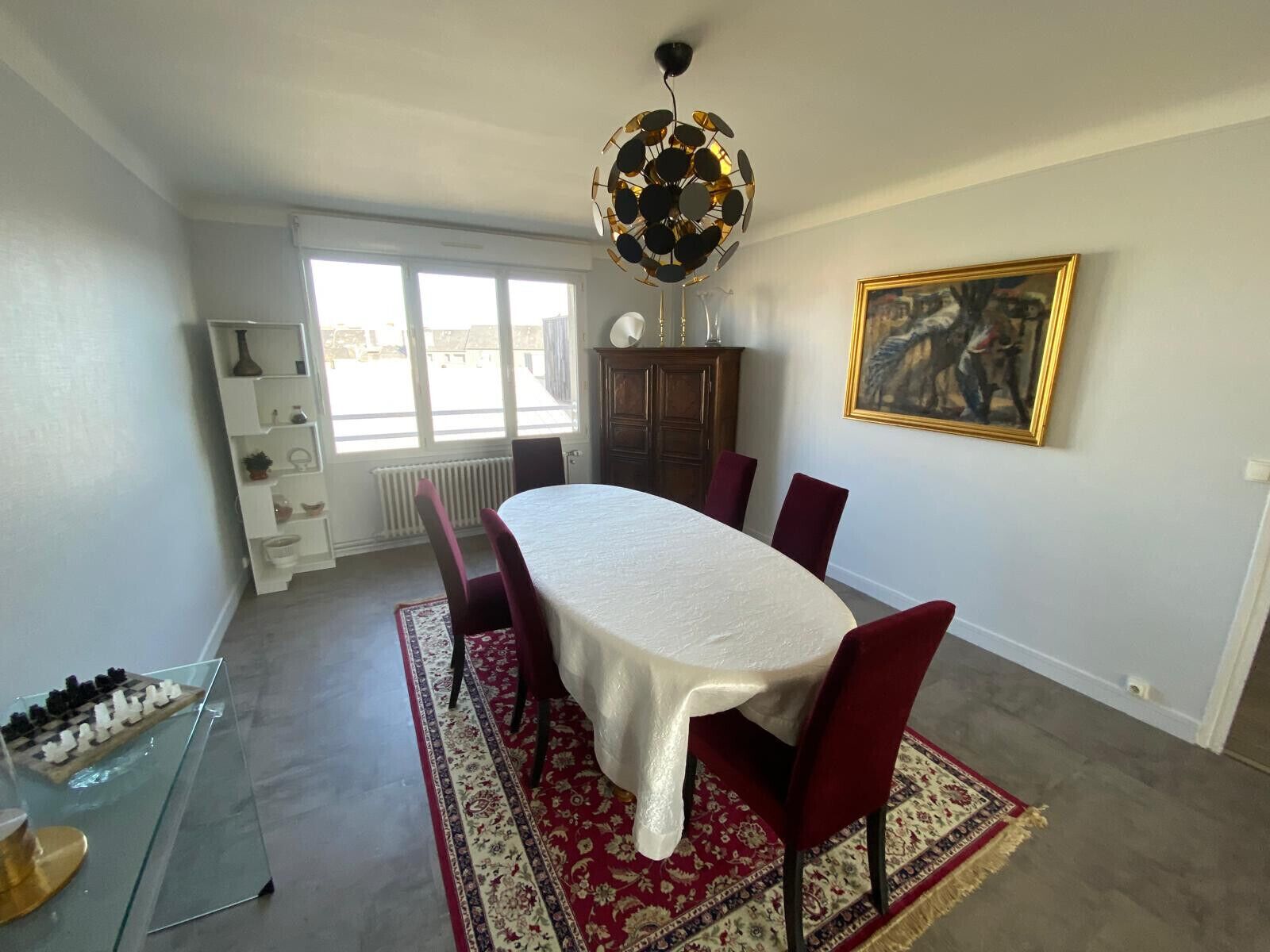 Un coin repas avec une table ronde, des chaises rouges, un tapis à motifs, un lustre et un tableau encadré accroché au mur.