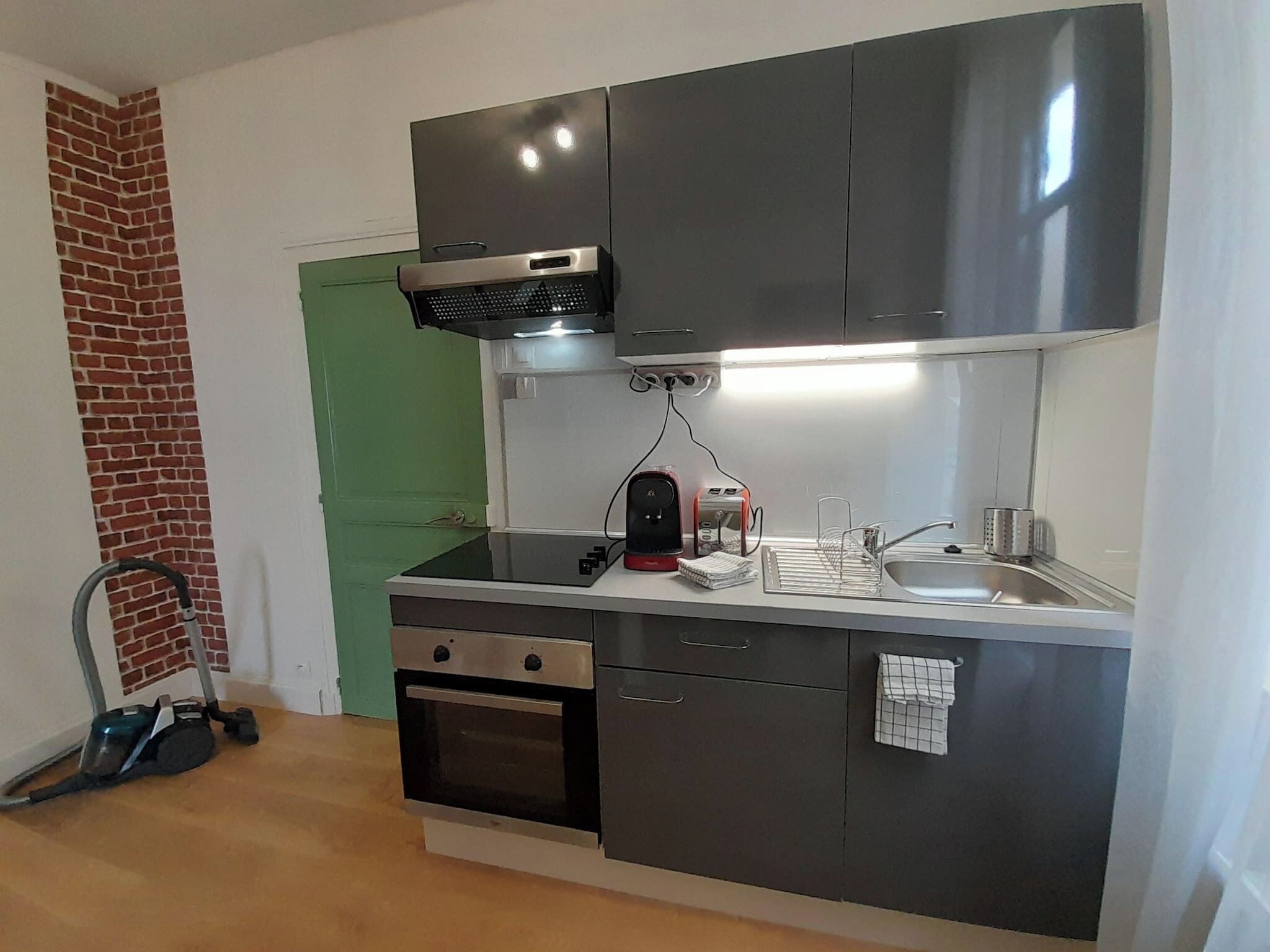A compact kitchen with a built-in oven, a microwave, and a sink. There is a vacuum cleaner in the corner, and a green door is visible in the background.