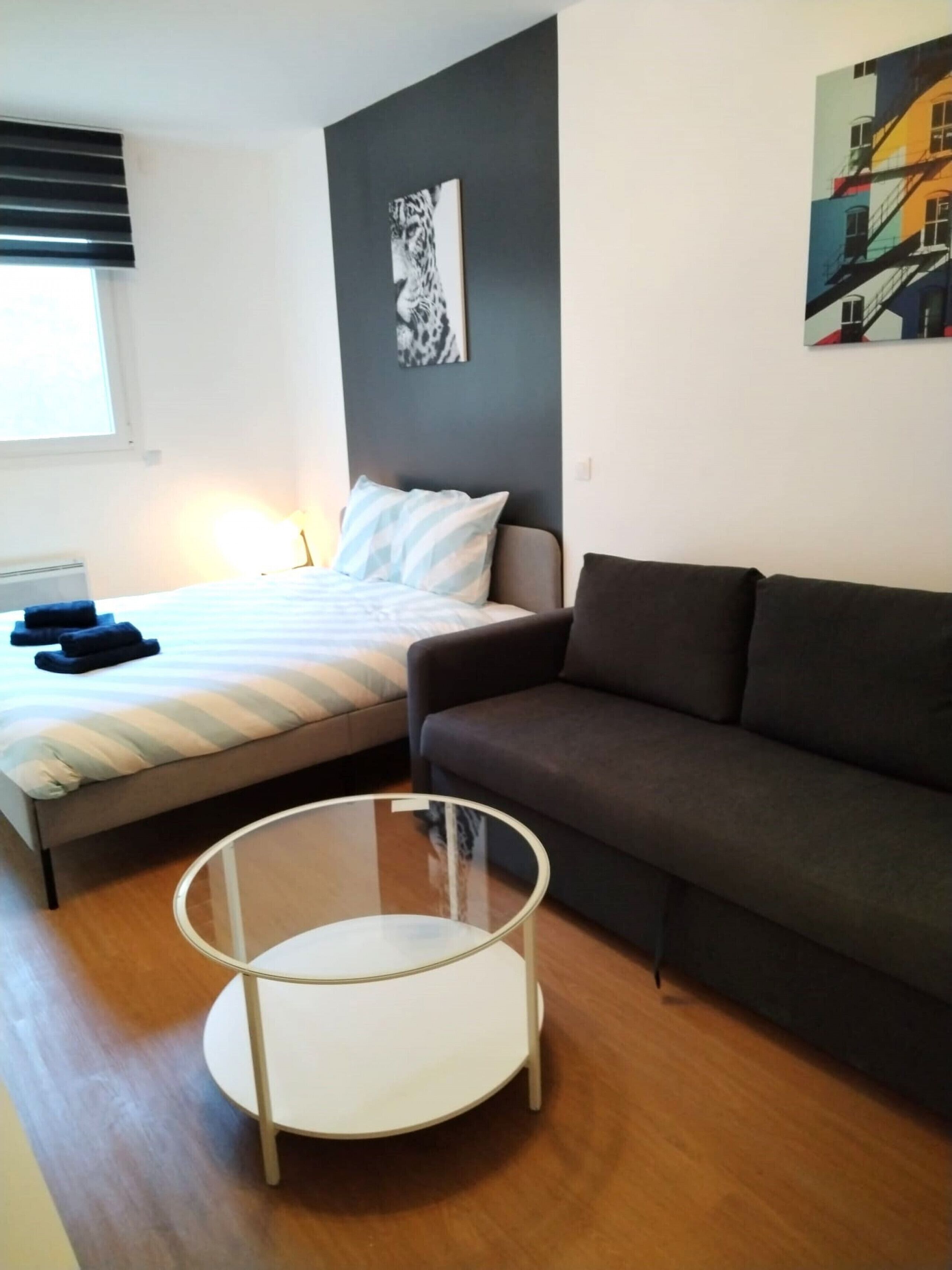 A modern bedroom with a bed, a grey sofa, a glass coffee table, and artwork on the walls.