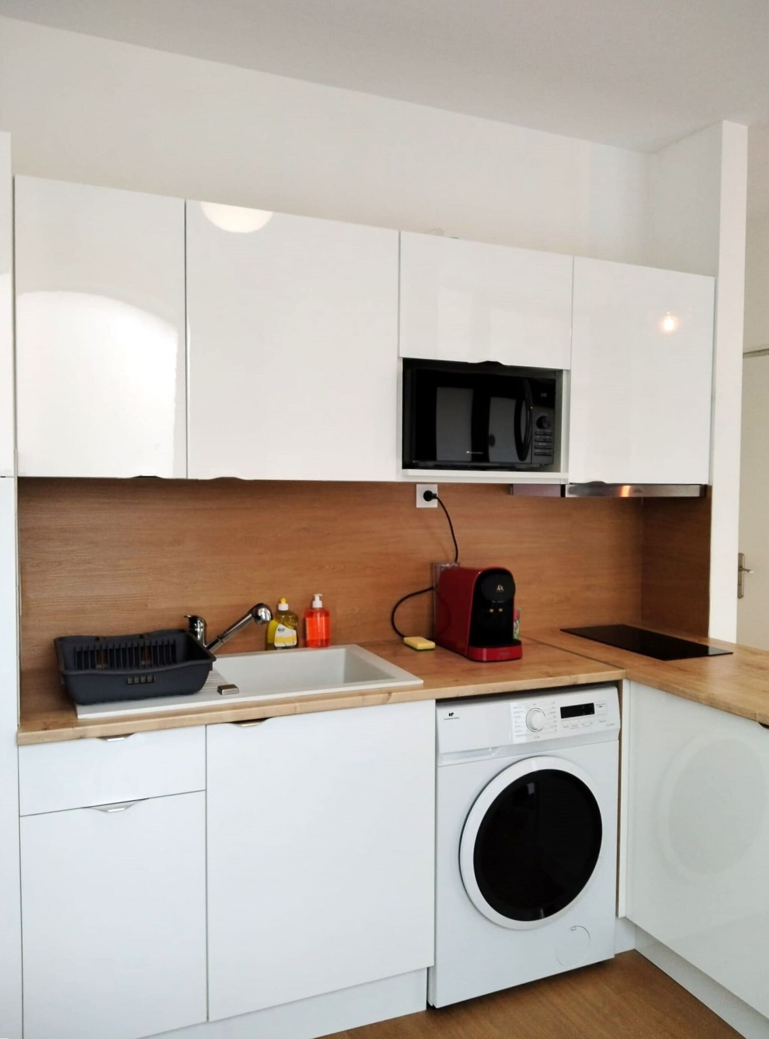 A compact kitchen with white cabinets, a microwave, a coffee maker, a sink, and a washing machine.