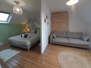 A bedroom with a bed, a grey sofa, a wooden floor, a hanging light fixture, and a wall with a brick pattern. - The workshop of dreams - 4 pers - wifi - air conditioning (Châteauroux)