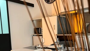 Ein moderner Essbereich mit minimalistischem Design, ein Tisch für zwei Personen gedeckt mit Gläsern und Tellern, eine Uhr an der Wand und eine Küchenzeile im Hintergrund.