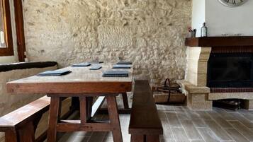 Un mur de pierres apparentes, une table en bois avec des bancs, et une cheminée surmontée d’une horloge.