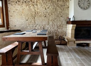 A rustic stone wall, a wooden table with benches, and a fireplace with a clock above it.