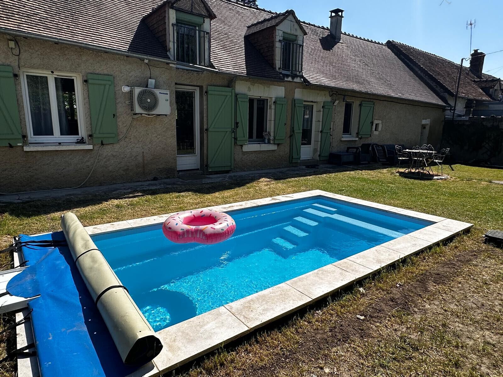 A swimming pool with a pink inflatable ring, a covered pool table, and a house with green shutters.