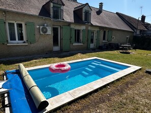 A swimming pool with a pink inflatable ring, a covered pool table, and a house with green shutters.