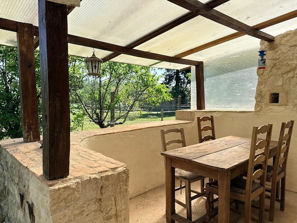 A covered patio with a wooden table and chairs, a hanging lantern, and a view of greenery.