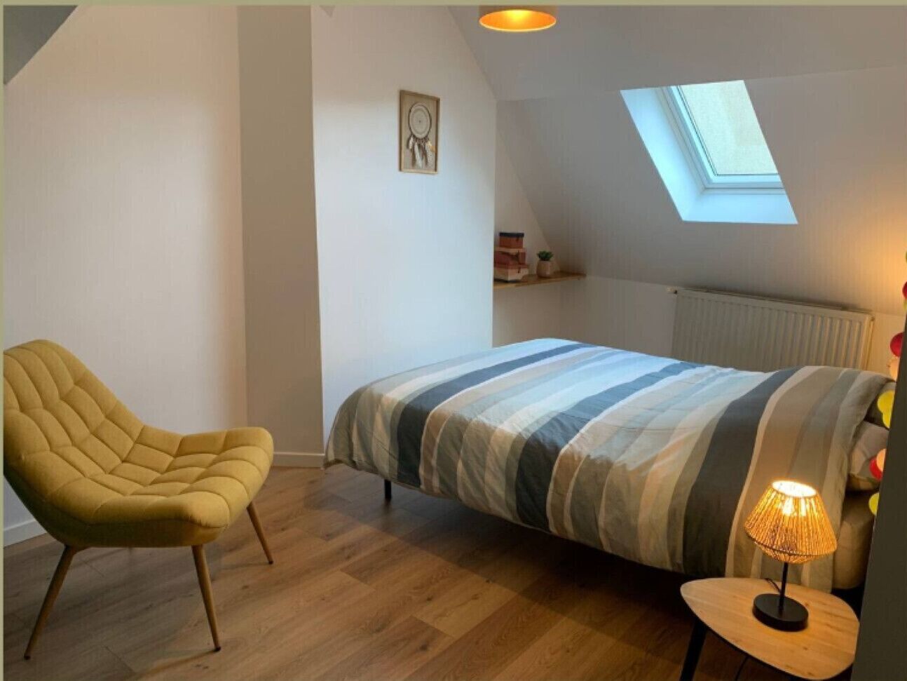 A bedroom with a bed, a chair, a lamp, a skylight, and a shelf with items.