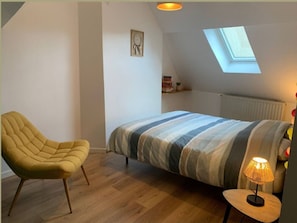 A bedroom with a bed, a chair, a lamp, a skylight, and a shelf with items. - Maison proche du lac-4 pers-wifi-cour (Châteauroux)