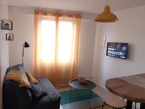 A living room with a sofa, a small round table, a television mounted on the wall, and a window with curtains.