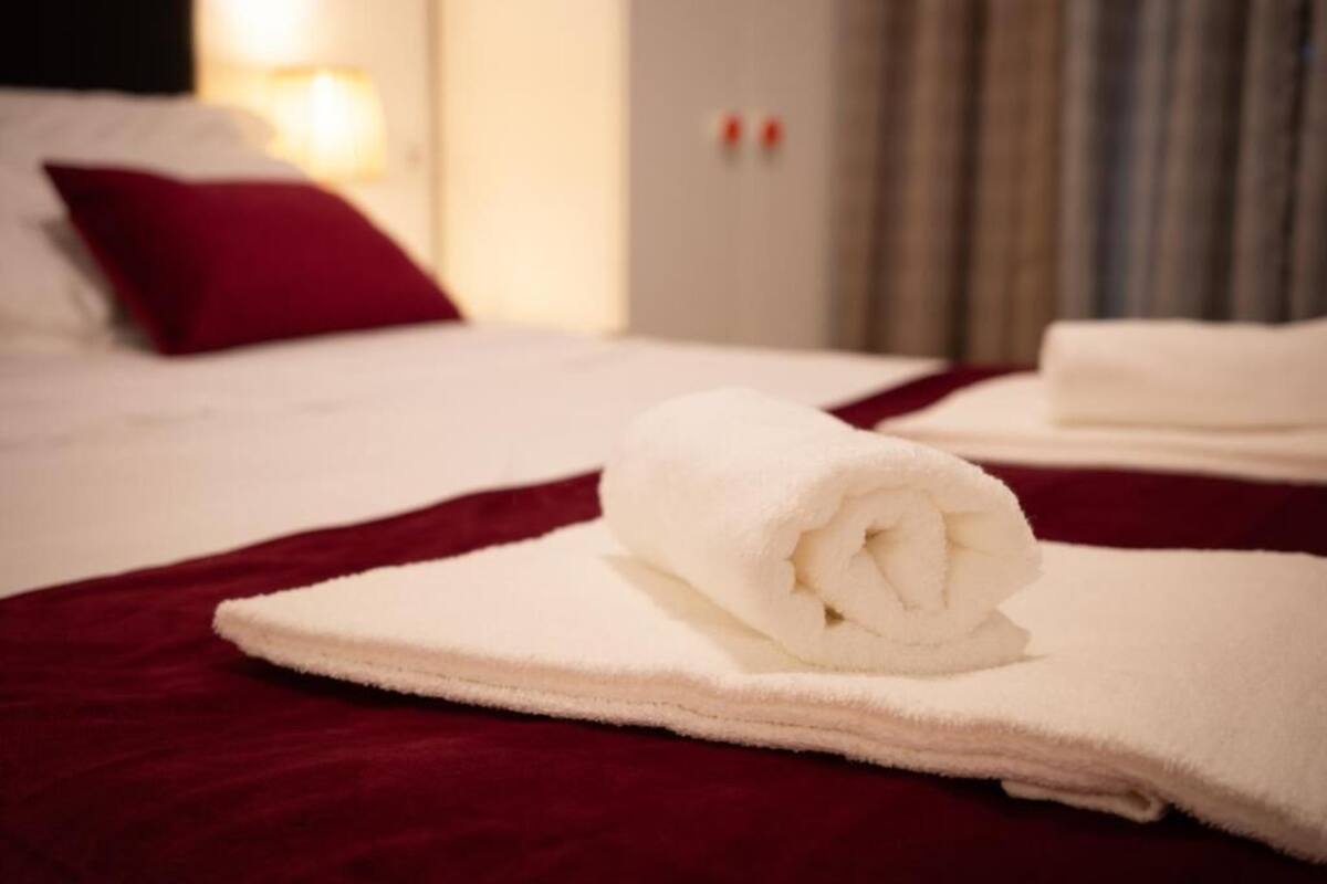 A neatly made bed with a red pillow and a rolled white towel on a red bedspread.