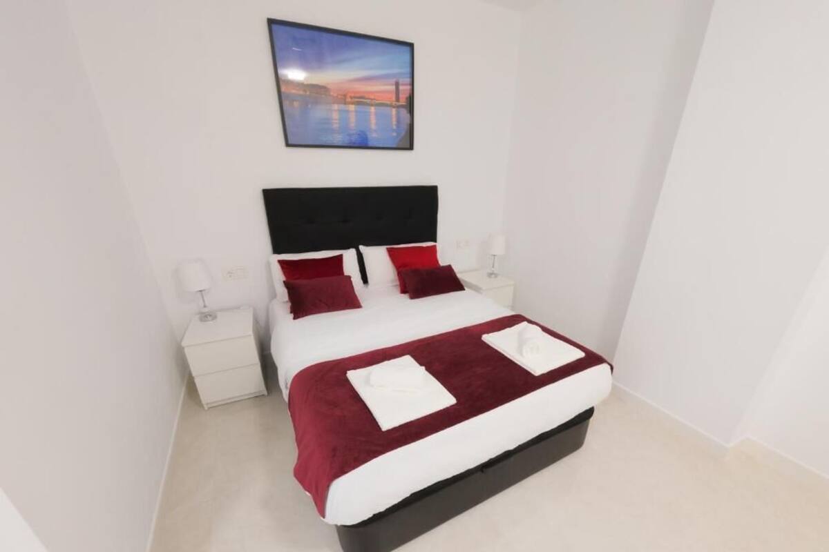 A neatly made bed with a red and white bedspread, two white pillows, and a black headboard. There is a white nightstand with a lamp on each side of the bed. A framed picture hangs above the bed.