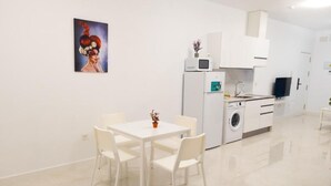 A modern kitchen with white cabinets, a microwave, and a washing machine. There is a dining table with chairs and a small potted plant.