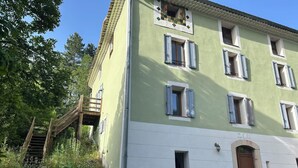 Un edificio de dos pisos con fachada verde y blanca, una escalera de madera que lleva a un balcón, y mobiliario exterior con sillas rosas.