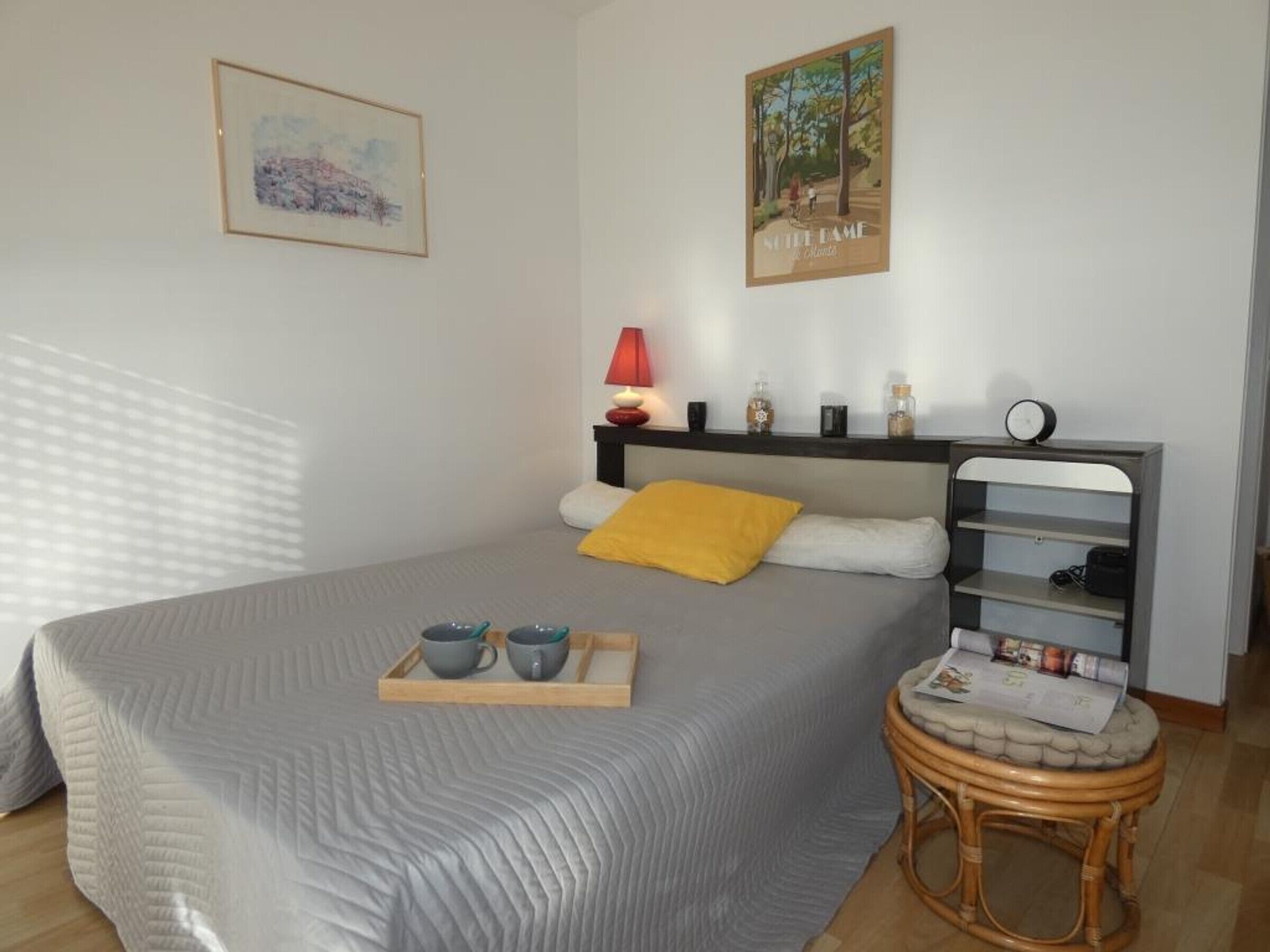 Une chambre à coucher avec un lit, une table de chevet avec une lampe, une petite table ronde sur laquelle se trouve un plateau avec des tasses, et un mur orné de deux tableaux encadrés.