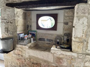 A stone-walled alcove with a small window, a metal bucket, a magazine rack, and a wireless charging station.