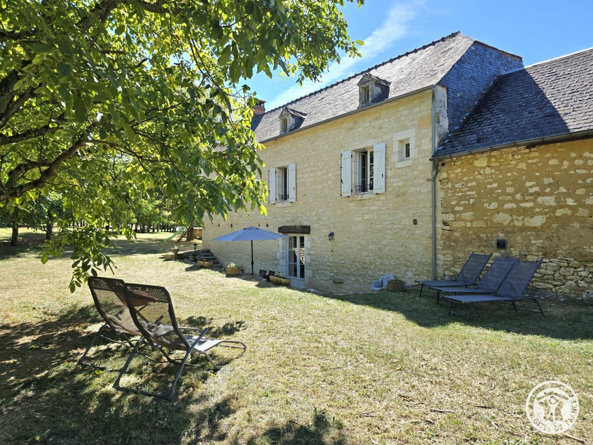 Un bâtiment en pierre avec un toit en pente, un patio couvert avec des chaises et un arbre qui offre de l’ombre.