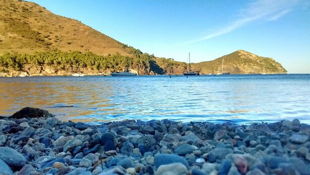 Una costa rocosa con aguas tranquilas, barcos y colinas al fondo.