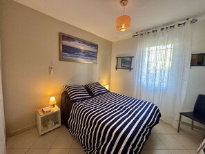Ein Schlafzimmer mit Bett, Nachttisch, Lampe, einem Gemälde, einem Sessel und einem Fenster mit Vorhängen.