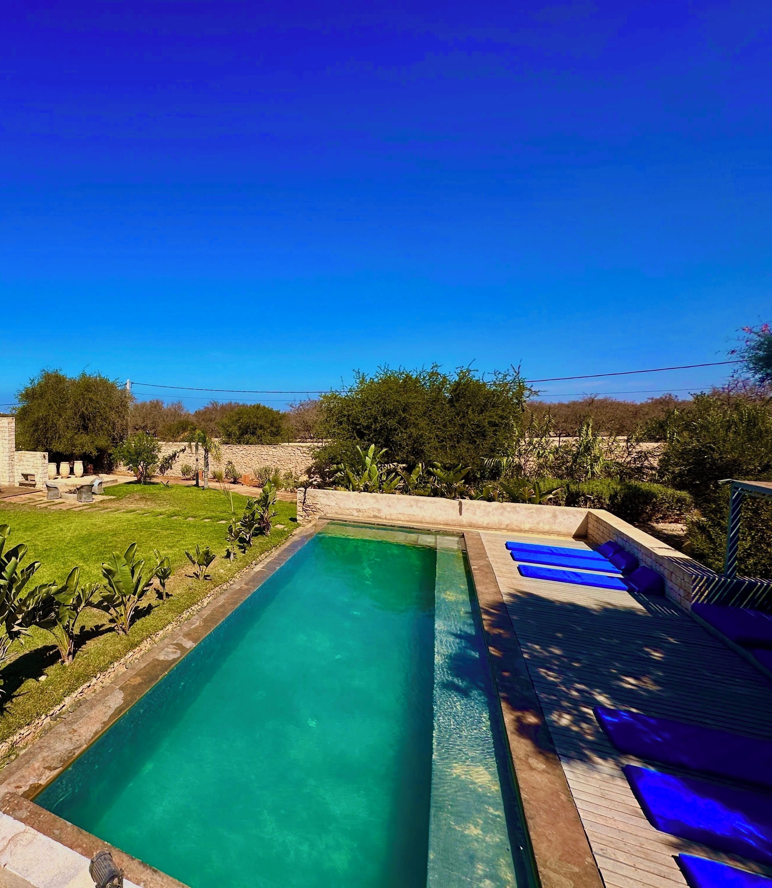 A rectangular swimming pool with clear blue water, surrounded by a stone wall and a wooden deck with blue lounge chairs.