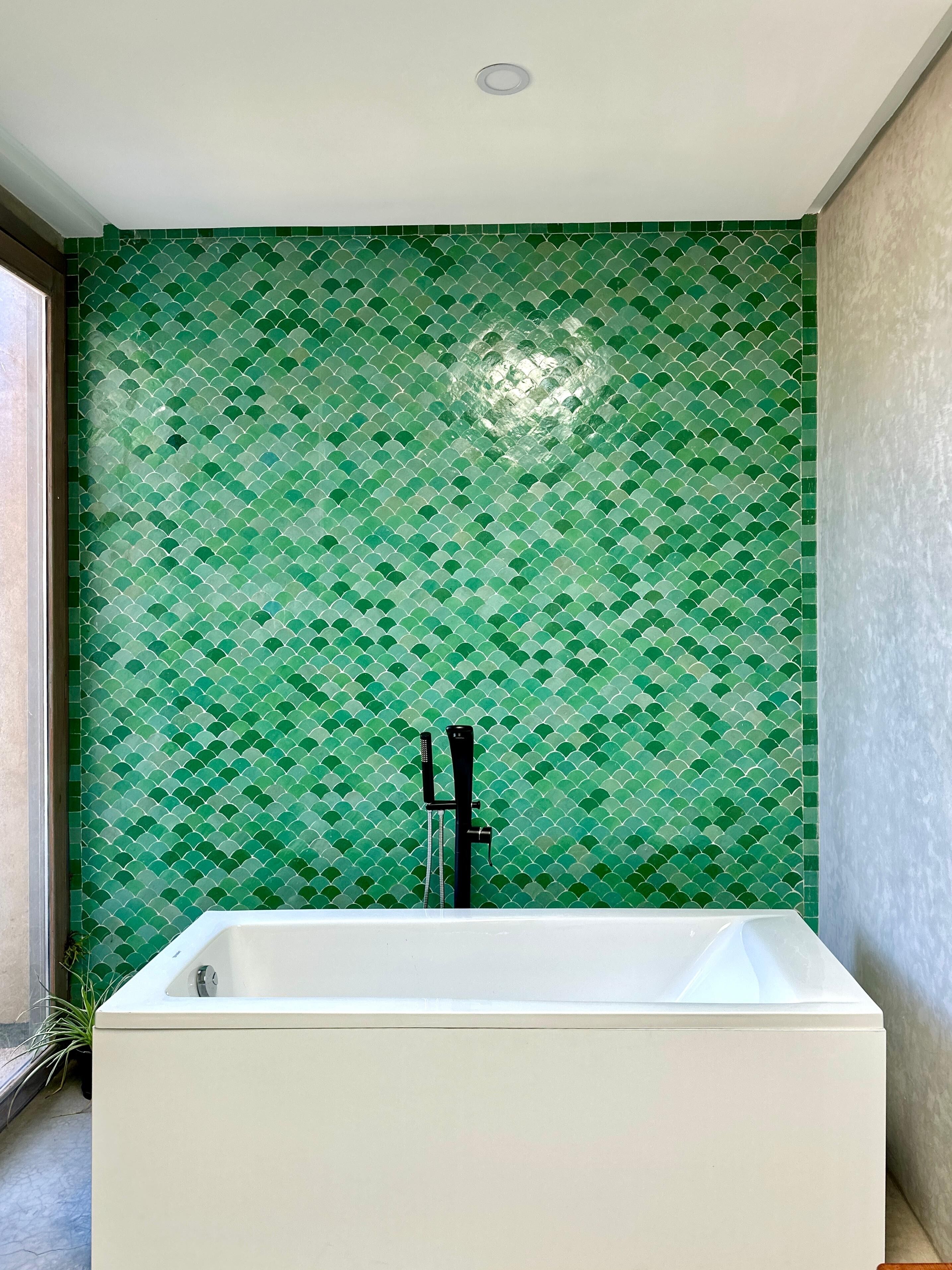 Casa de banho com parede em mosaico verde, uma banheira branca e uma torneira preta.