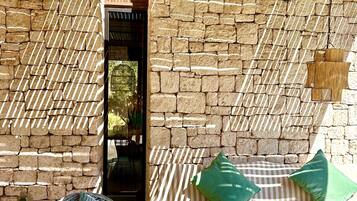 Una habitación con pared de piedra, una silla de mimbre, un sofá a rayas y una mesa con una maceta roja.