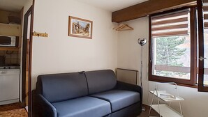 Un espace de vie compact comprenant un canapé, une petite kitchenette, une fenêtre avec des stores et un tableau au mur.
