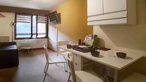Une pièce compacte comprenant un coin cuisine, une table à manger avec des chaises, un petit canapé et une fenêtre avec des stores.