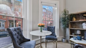 A corner room with a round table, two chairs, a bookshelf, and a large window with a view of buildings. - 2 Bed Luxury Condo in South End near Restaurant Row, (Boston)