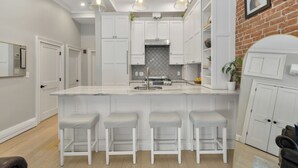 A modern kitchen with white cabinetry, a central island with stools, pendant lighting, and a brick accent wall. - 2 Bed Luxury Condo in South End near Restaurant Row, (Boston)
