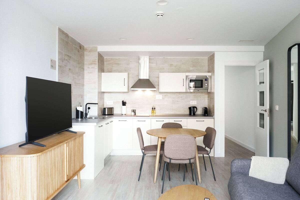 Una cocina moderna con armarios blancos, un microondas y una cafetera. Hay una zona de comedor con una mesa y sillas.