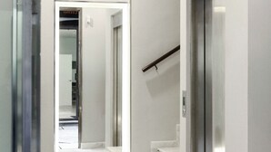 Un ascensor moderno con detalles metálicos, una escalera con escalones de mármol y una puerta de cristal que da a una habitación.
