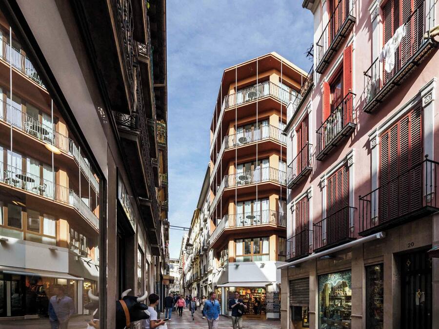 A narrow street flanked by multi-story buildings with balconies, shops on the ground floor, and a clear sky above.