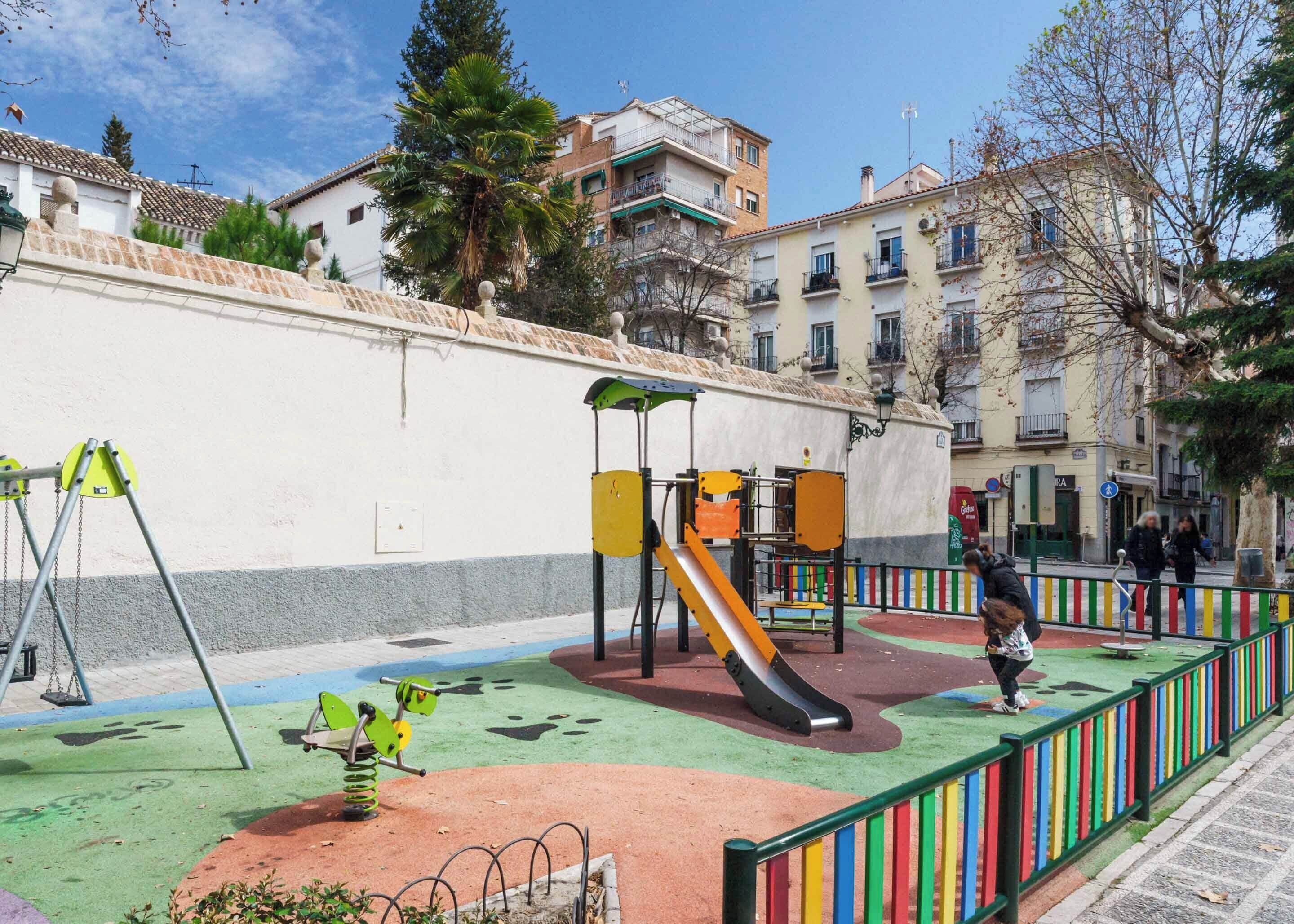 A playground with swings, a slide, and climbing structures, surrounded by a low wall and colorful railings.