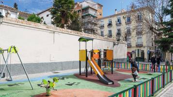 A playground with swings, a slide, and climbing structures, surrounded by a low wall and colorful railings.