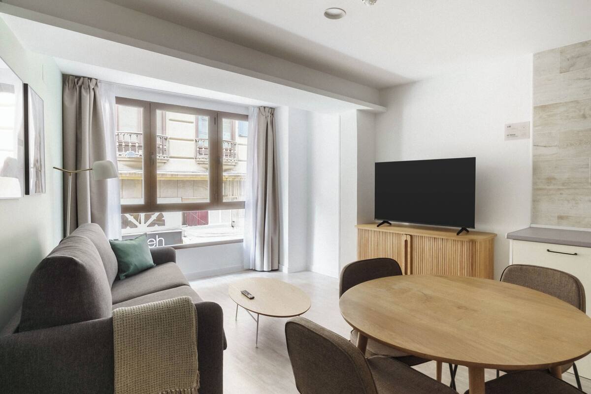 Una sala de estar moderna con un sofá, una mesa de centro, un televisor sobre un mueble de madera y una mesa de comedor con sillas.