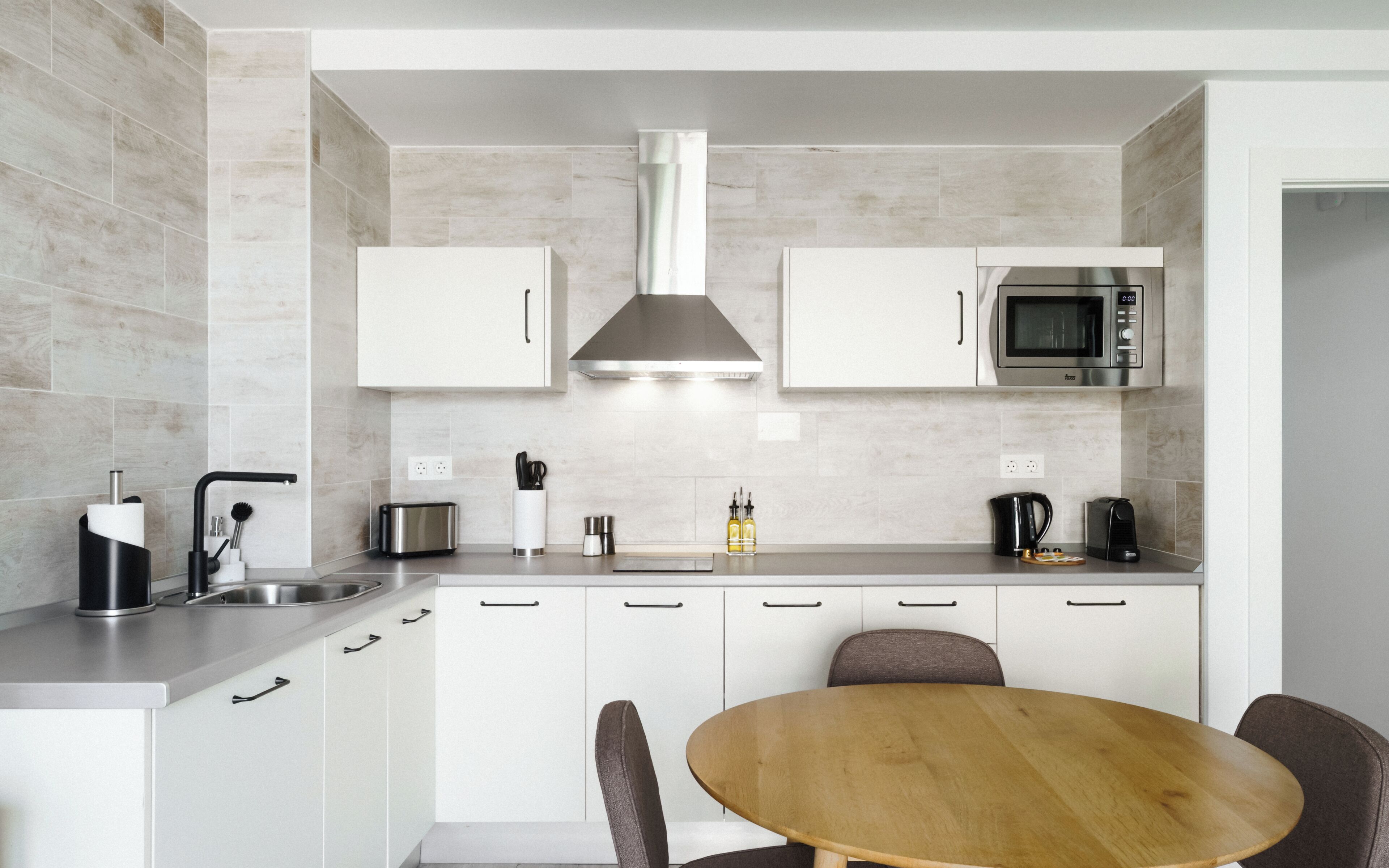 Una cocina moderna con armarios blancos, una campana extractora de acero inoxidable, un microondas y una mesa de comedor de madera con sillas.