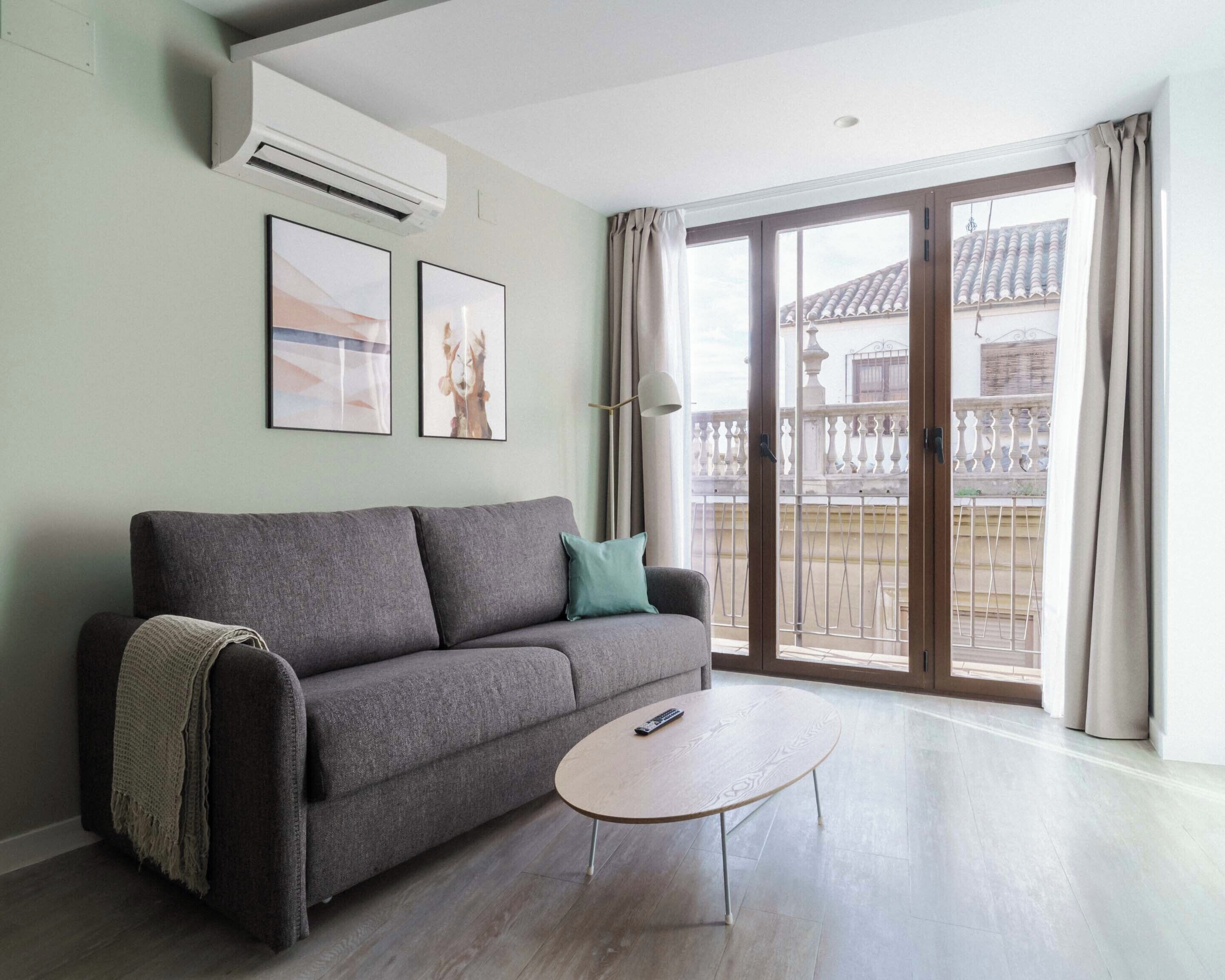 A living room with a grey sofa, a small round table, and a balcony with a view of buildings.