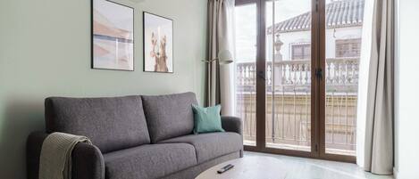A living room with a grey sofa, a small round table, and a balcony with a view of buildings.