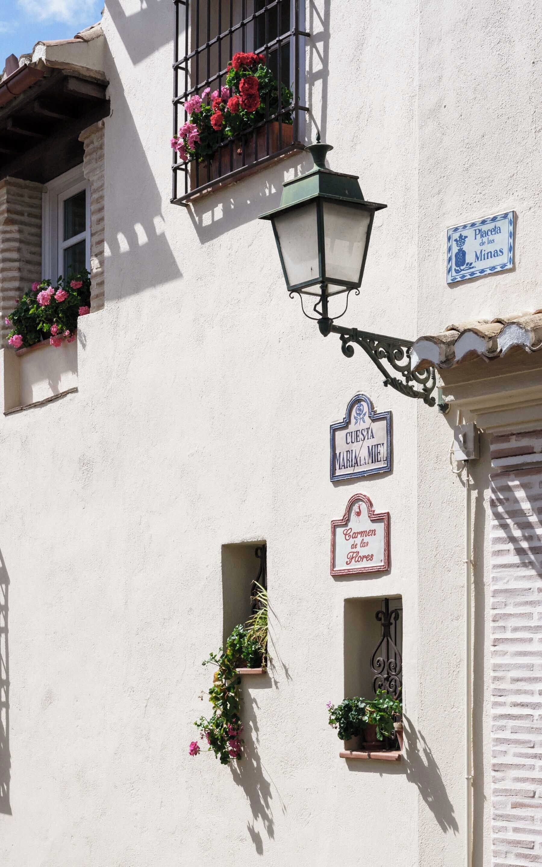 A building with a white facade, a green street lamp, and signs indicating 'CUESTA MARIA LA MET' and 'Carme de las Flores'.
